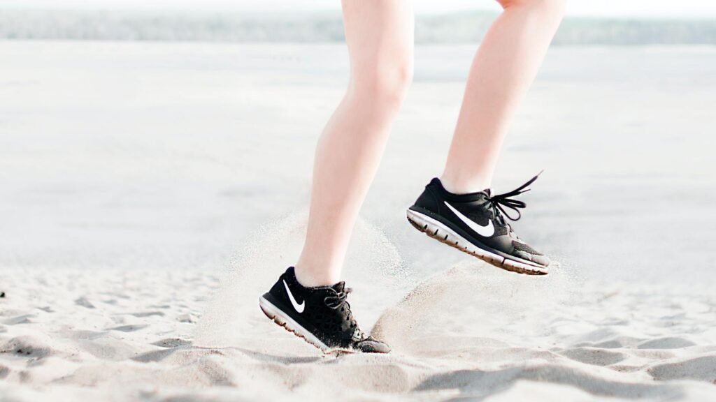 Une femme en chaussures de course Nike et aux jambes épilées, sautant dans le sable.