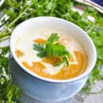 roasted butternut squash soup in a blue china bowl with green cilantro around it and a splash of cream on top. Sitting on a silver tray and blue tablecloth.
