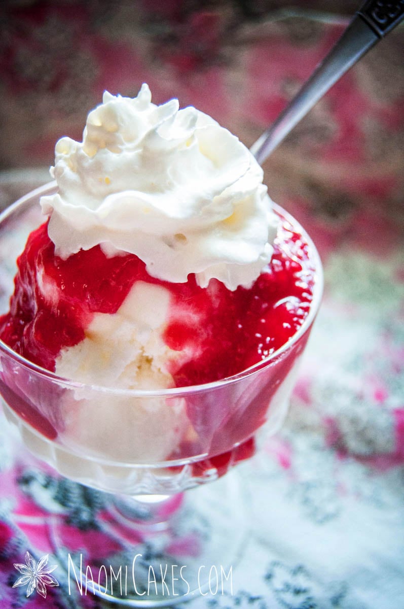 close up photo of a glass stand dessert cup with ice cream, raspberry sauce and whipped cream and a spoon