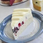 a slice of lemon mousse cake with fresh berries in it on a blue china plate with two cakes in the background