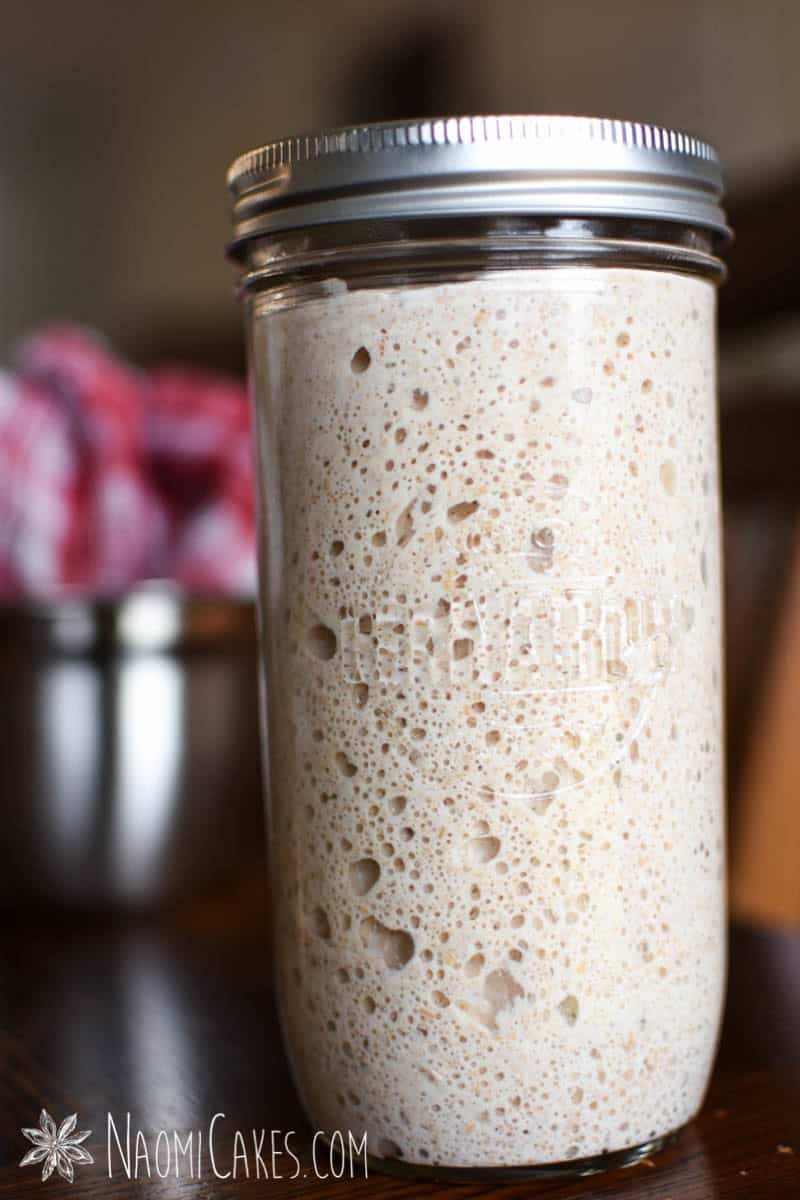 close up of a jar of whole wheat sourdough starter on a wooden table with bowls and tea towels in the background