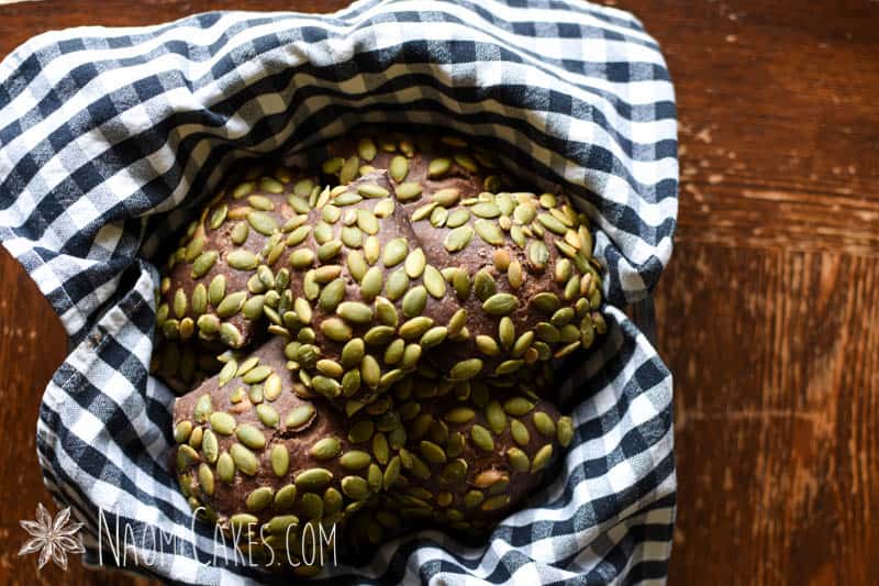 baked dark rye pumpkinseed covered rolls in a bowl with a checked cloth