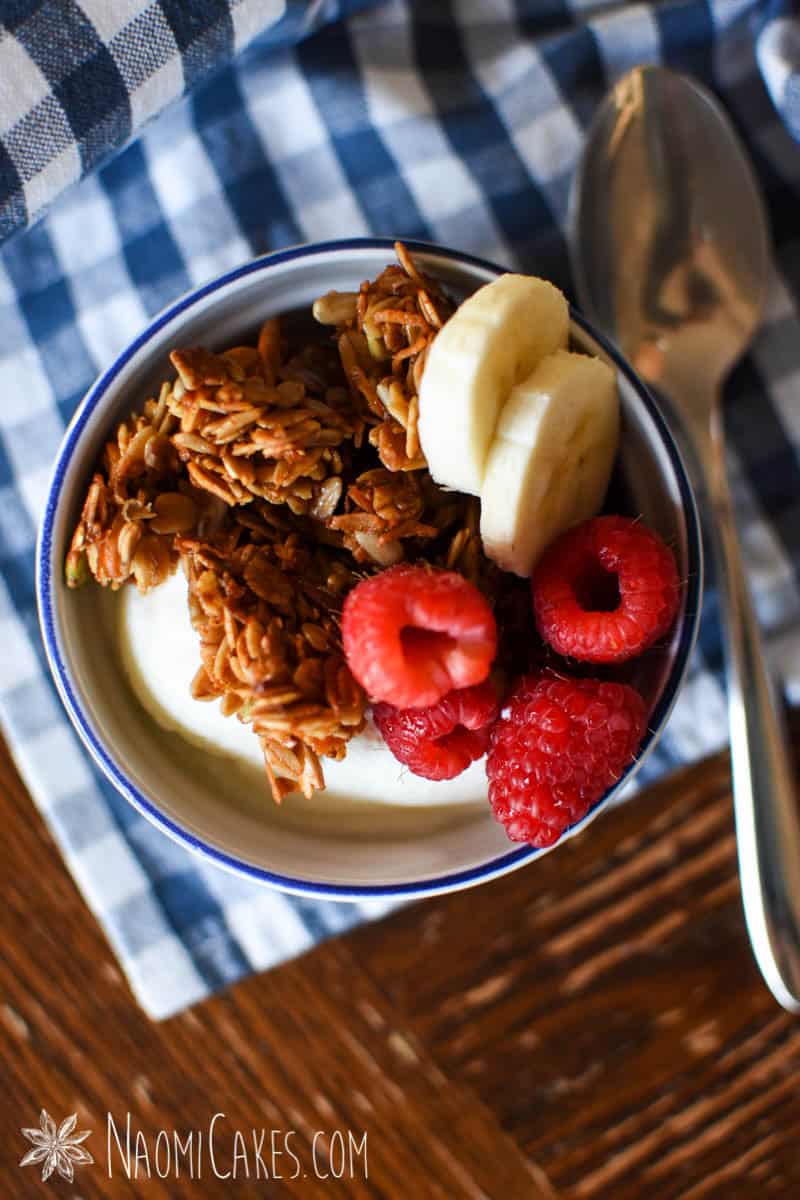 top view of a ramekin with yogurt topped with granola, raspberries and banana slices on a blue gingham cloth