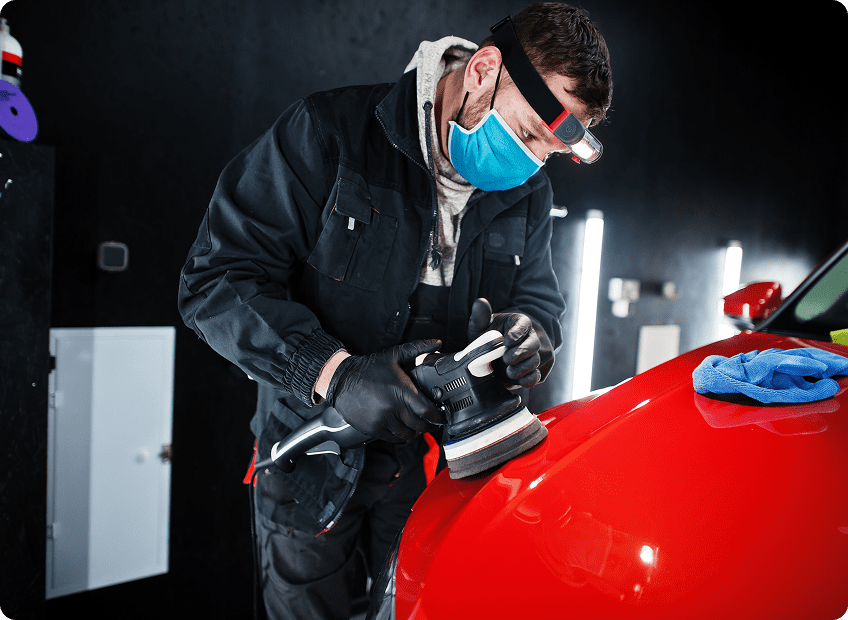 A man wearing a mask, gloves, and a headlamp uses a polishing tool on the shiny red hood of a car in a well-lit garage, with a blue cloth nearby.
