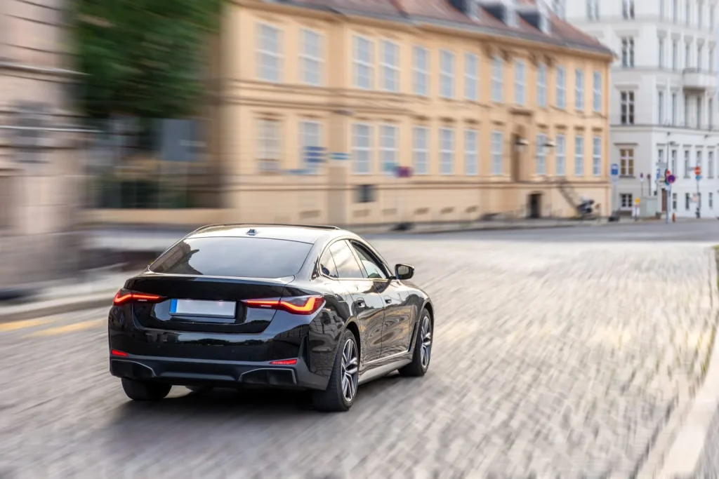 A sleek black sedan drives quickly down a cobblestone street in a European city, with historic buildings and blurred motion in the background.
