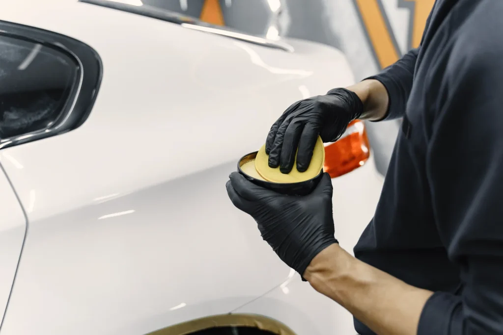 A person wearing black gloves applies car wax with a yellow applicator pad to the side of a white vehicle.
