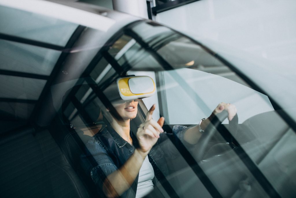 A person sitting in the driver’s seat of a car wears a virtual reality headset and touches the air with one hand, as seen through the windshield.