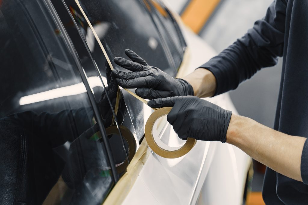 A person wearing black gloves applies masking tape along the edge of a car window, preparing the vehicle for painting or detailing.