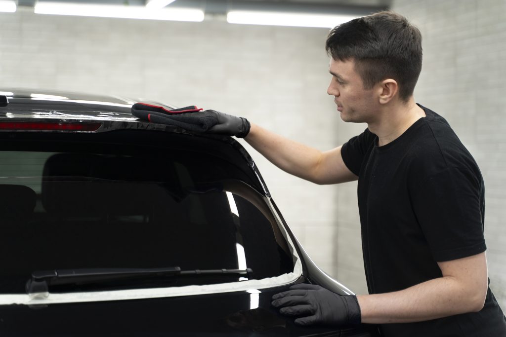 A man wearing black gloves and a black t-shirt is polishing the rear window area of a black car in a well-lit garage or workshop. White tape borders the window, indicating ongoing detailing work.