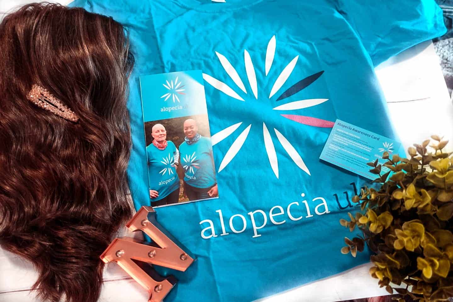 Alopecia Awareness Month flat lay with Alopecia UK t-shirt, wig and plant