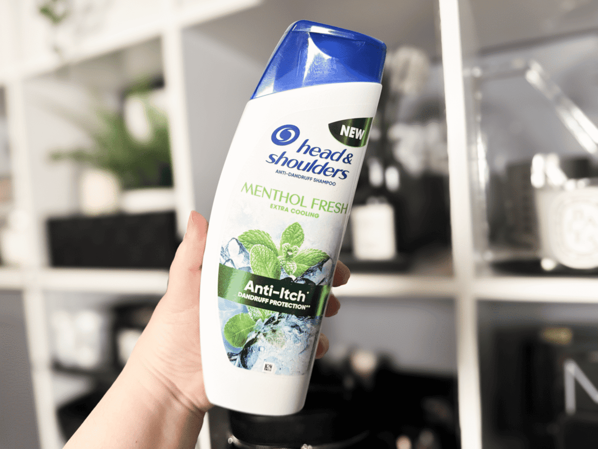holding up a bottle of the head and shoulders menthol shampoo infront of a blurred white shelf background