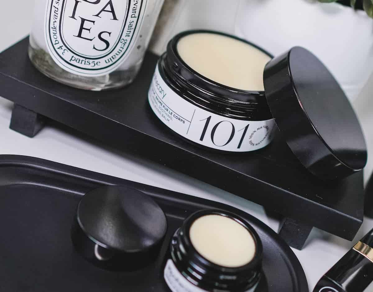 product shot of both the sos body balm and the lip balm with their lids twisted off and slanted to the side of the product. Both products appeasr off-white in color and appear balmy in texture.