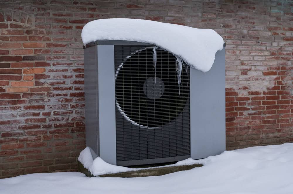 An air conditioning unit battles the winter chill, covered in a blanket of snow against a rustic brick wall.