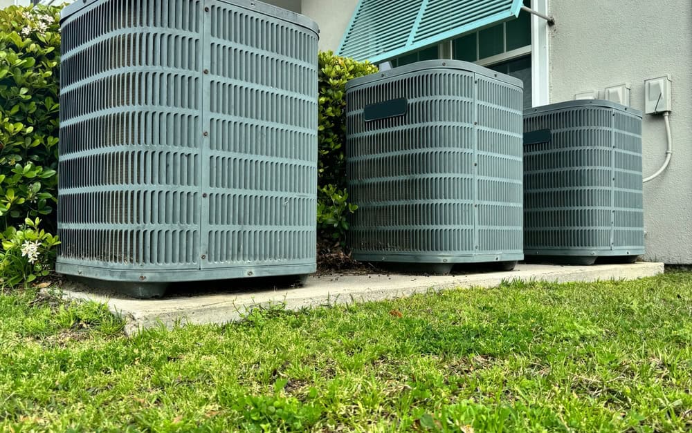 Three air conditioning units quietly humming in a lush green backyard.