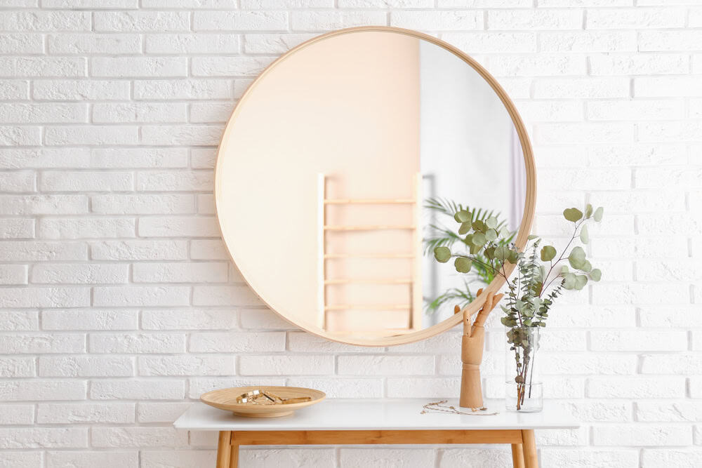 A minimalist white brick wall with a round mirror reflecting a wooden hand sculpture and eucalyptus leaves on a sleek wooden console table.