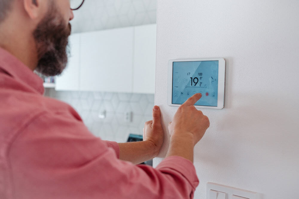 A man adjusts the digital thermostat on a smart home control panel in a modern kitchen.