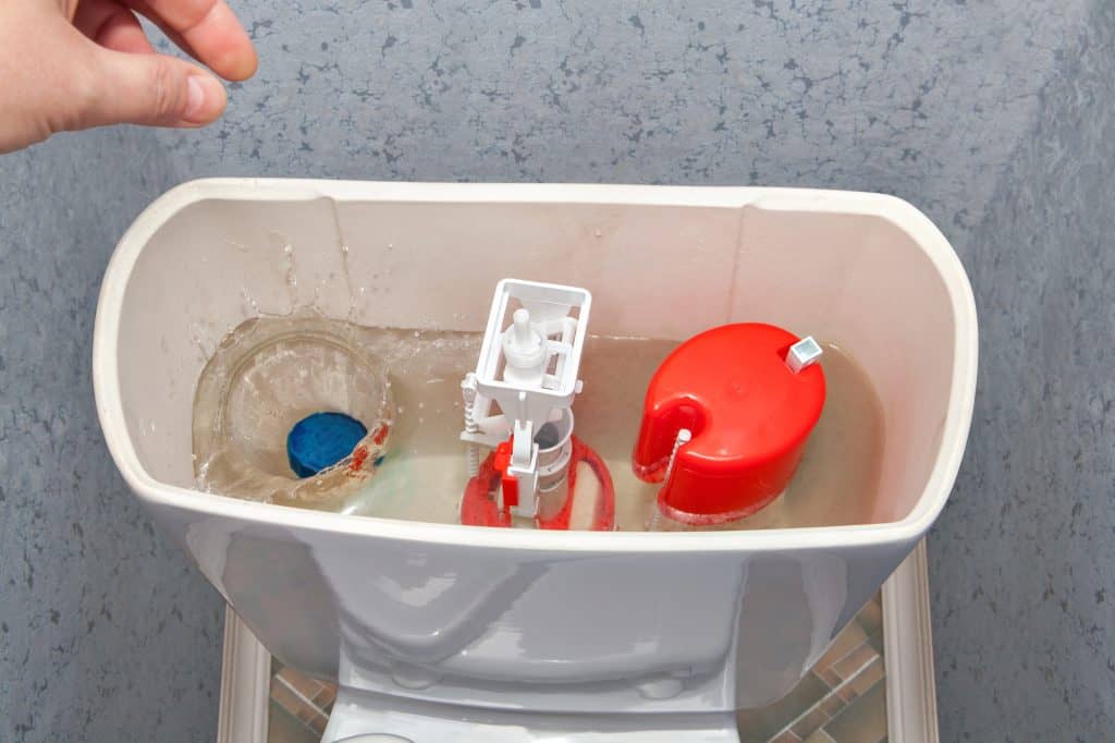 A hand drops a blue object into a toilet tank, revealing the inner workings including the fill valve and flush valve.