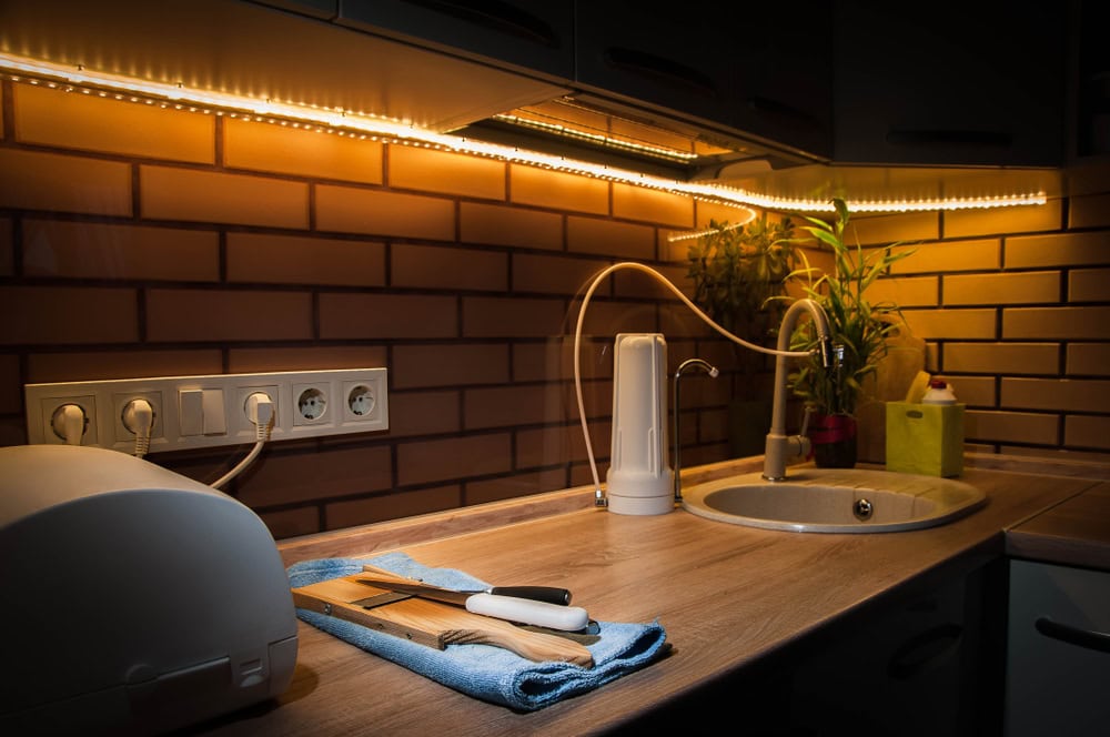 Modern kitchen workspace illuminated by warm LED strip lighting, featuring a wooden countertop, water filter, sink, and under-cabinet power outlets against exposed brick walls.