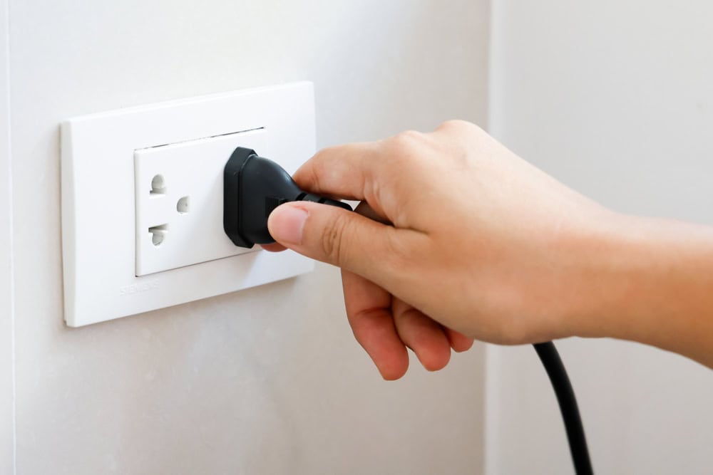 Plugging in: A hand connects a black power cord to a white electrical outlet mounted on the wall.