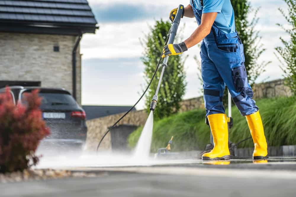 A worker in yellow boots power washes a driveway outside a modern house on a sunny day.