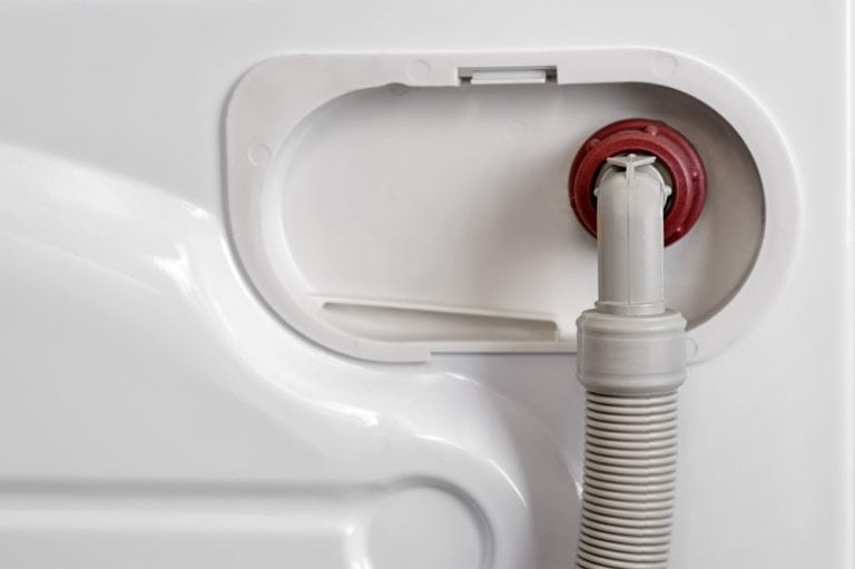 Inside a washing machine drain hose connection point, showing a white corrugated drain hose with a red seal mounted in a recessed access panel.