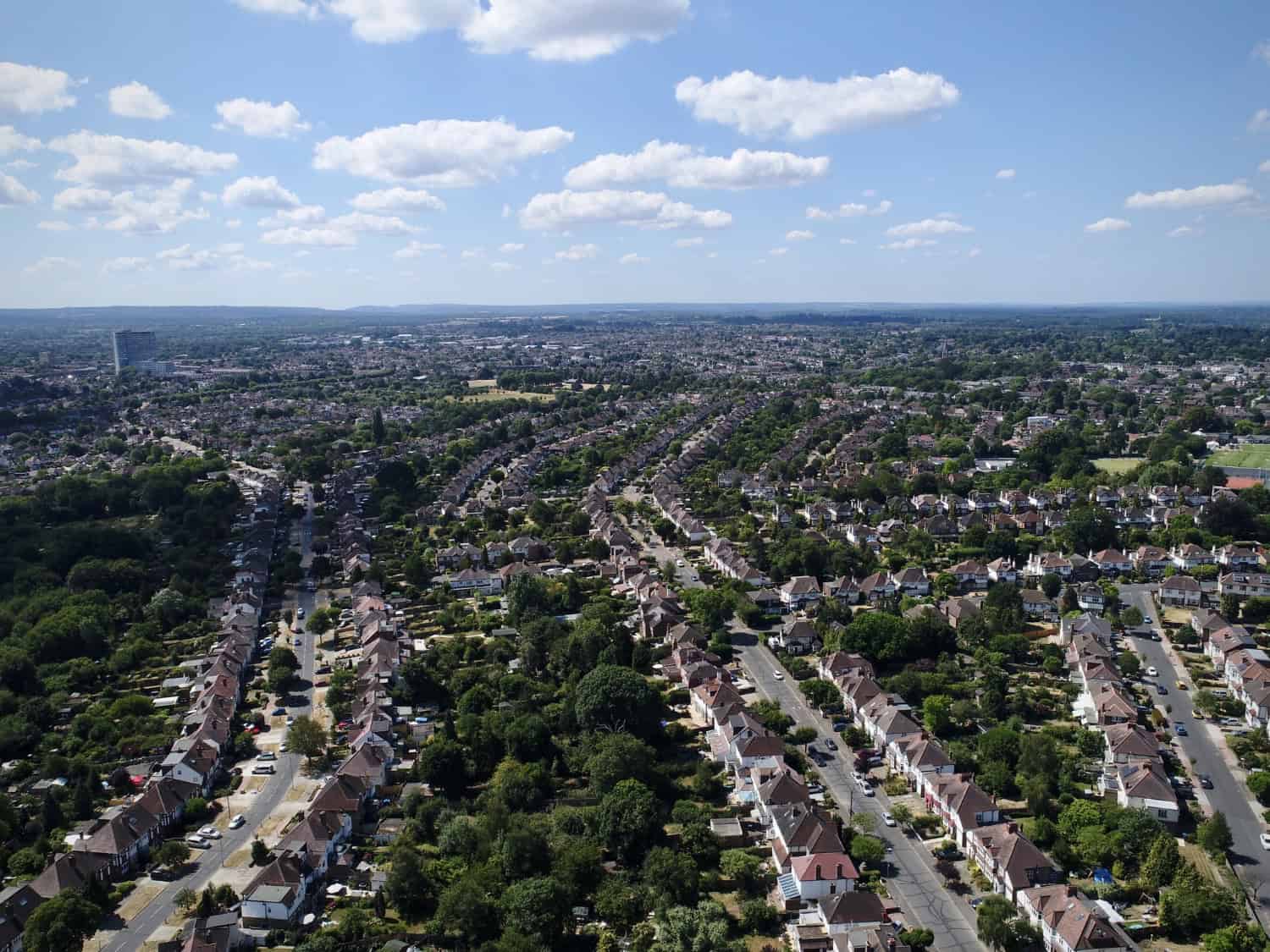 Aerial view of a suburban neighborhood with neat rows of semi-detached houses, tree-lined streets, and sprawling residential areas stretching toward the horizon under a cloud-dotted summer sky.
