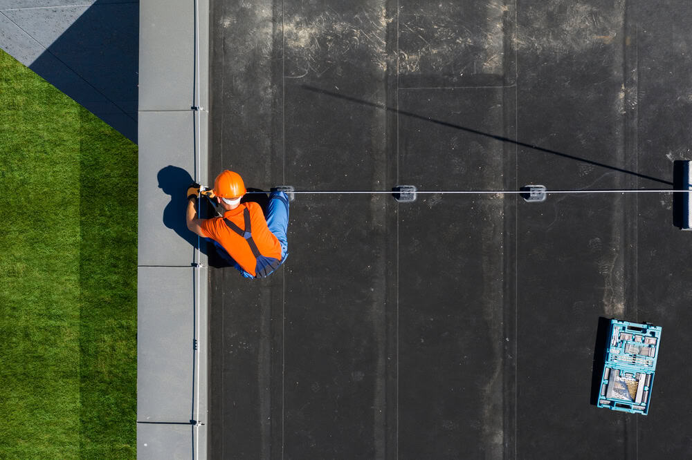 A worker in an orange helmet and vest secures safety lines on a rooftop, with a green field visible below.