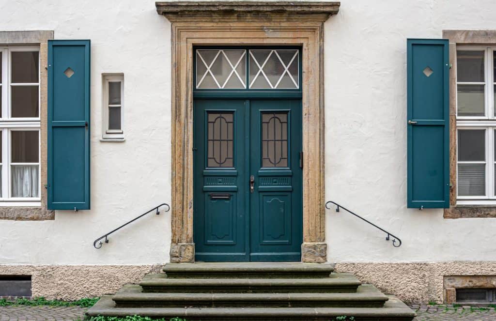 A charming teal door framed by elegant stonework, flanked by matching shutters on a quaint white building.