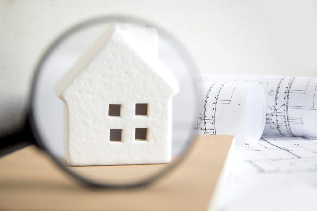 A magnifying glass focuses on a small white house model beside blueprints, highlighting real estate evaluation and planning.