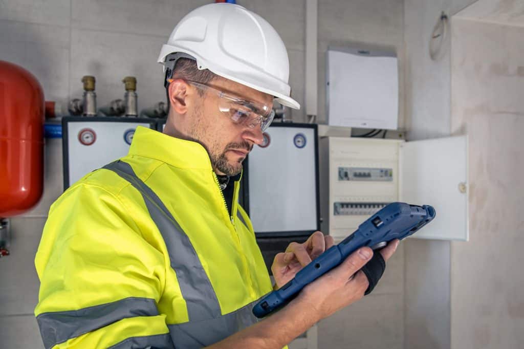 Safety-equipped maintenance worker reviews data on a handheld device while inspecting industrial equipment controls