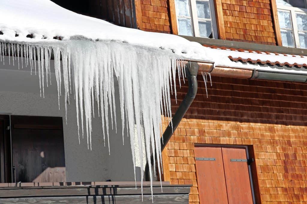 Dramatic icicles dangle from a snow-covered roof, contrasting against cedar shingles and copper gutters of a rustic building