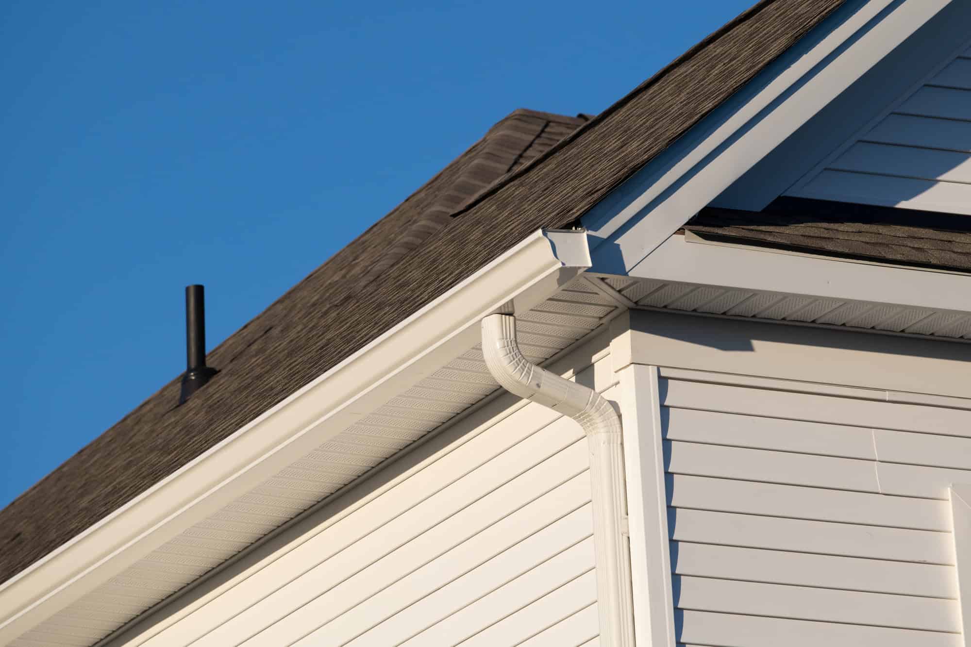 White rain gutter and downspout detail against weathered gray shingles and vinyl siding under bright blue skies