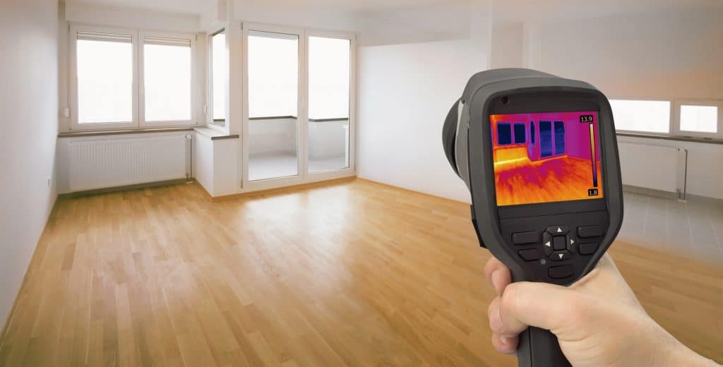 Thermal imaging camera reveals heat patterns in an empty apartment room with hardwood floors and large windows, highlighting temperature variations and potential insulation spots.