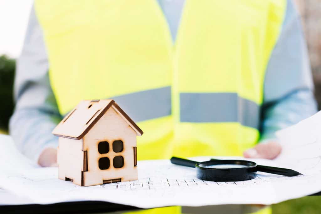 Building inspector examining plans with wooden model house and magnifying glass