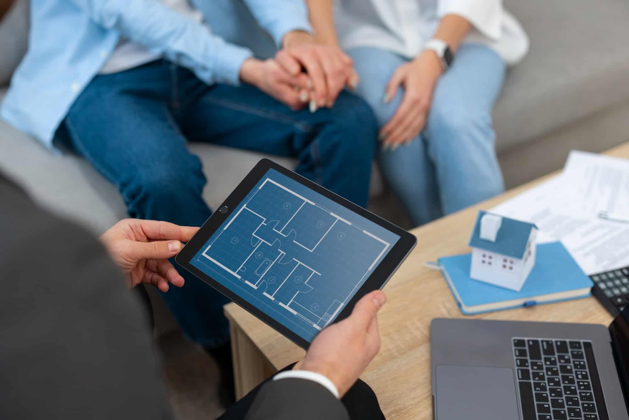 Real estate professional reviewing digital floor plans with clients, alongside a miniature house model and laptop during a consultation