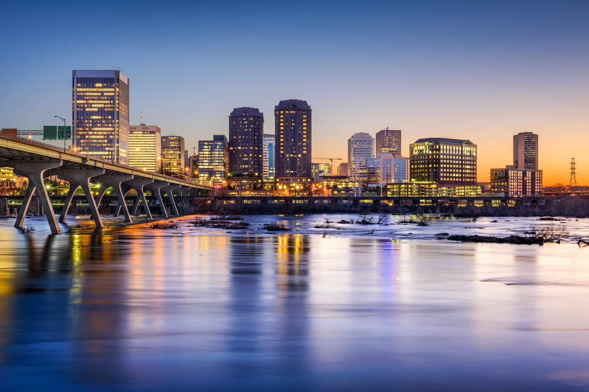 Richmond, Virginia skyline at dusk, with the James River reflecting the city lights and the distinctive arch supports of the Manchester Bridge spanning the partially frozen waters.