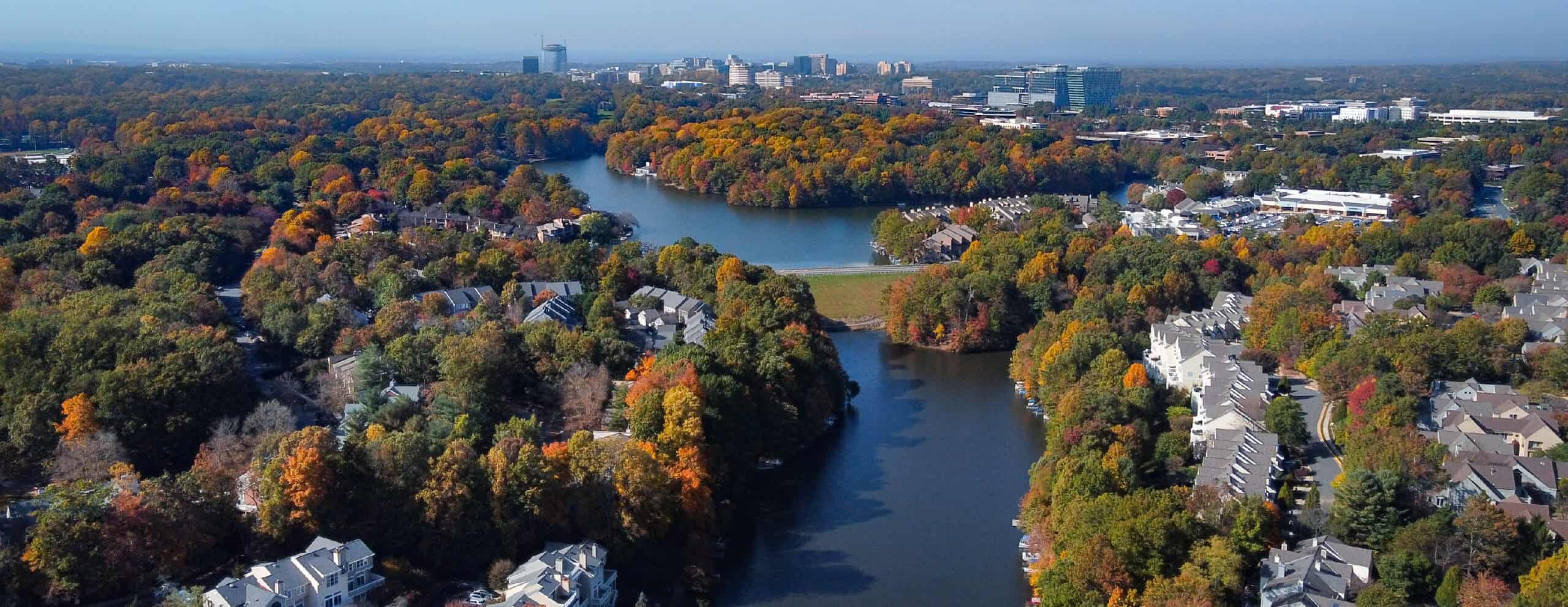 An aerial view of homes and townhouses along a wooded lake in Fairfax County, Virginia, with a distant downtown skyline.