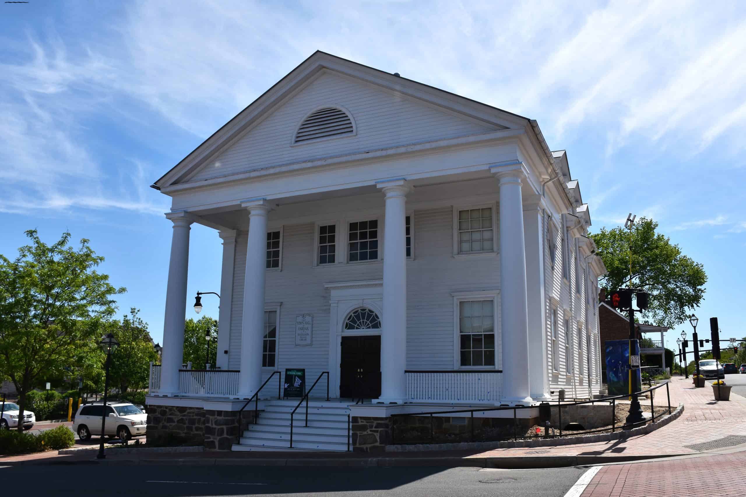 The white Fairfax Town Hall building with tall columns and a triangular pediment under a blue sky.
