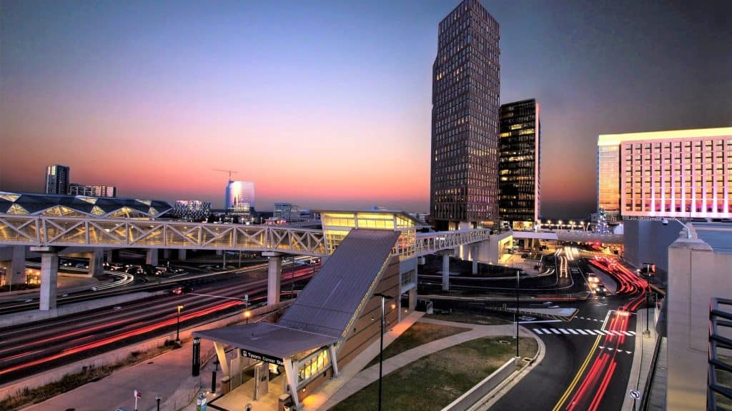 An elevated Metro station, pedestrian bridge and office towers illuminated at dusk.