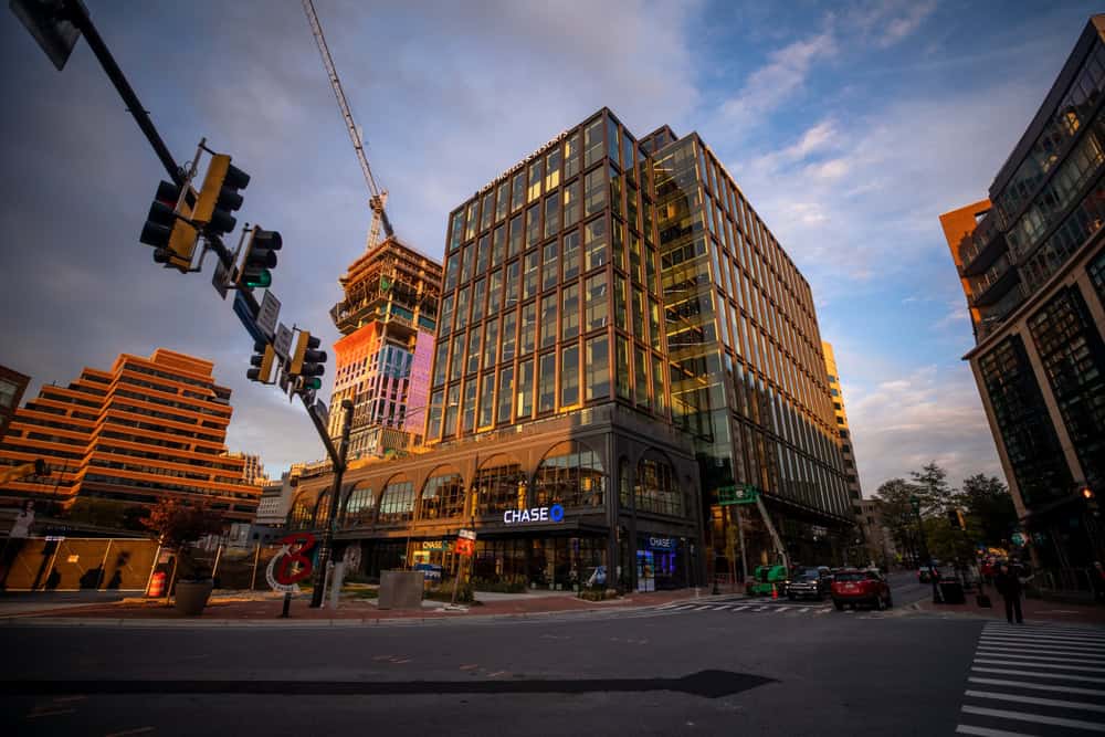 A glass and brick office tower under construction at dusk, with a Chase bank at street level and traffic signals in the foreground of a downtown intersection