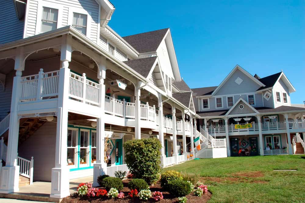 A row of white two-story buildings with wraparound porches, teal-trimmed shop windows and landscaped flower beds along the walkway