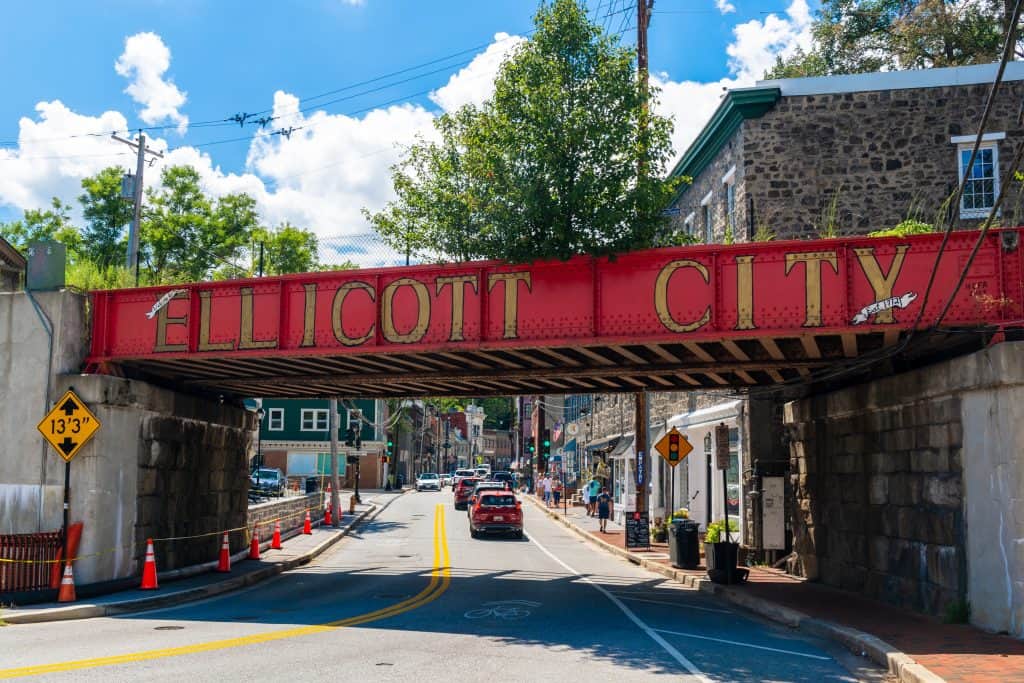 A red railroad bridge lettered Ellicott City spans Main Street's stone buildings and shops in Howard County, Maryland.