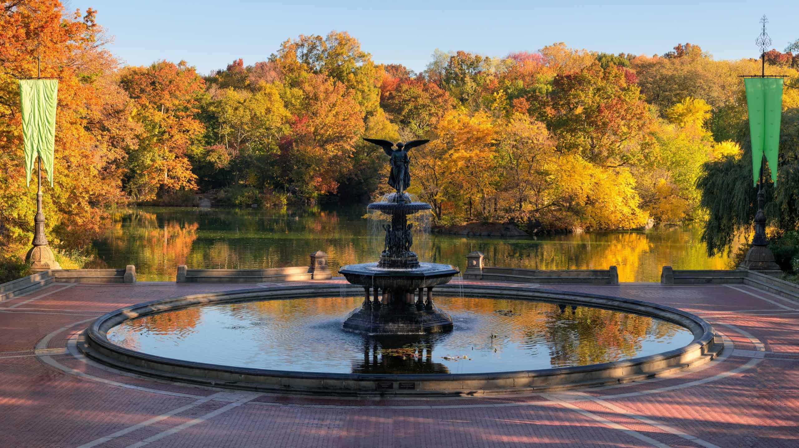 A fountain beside a lake framed by trees with autumn foliage.