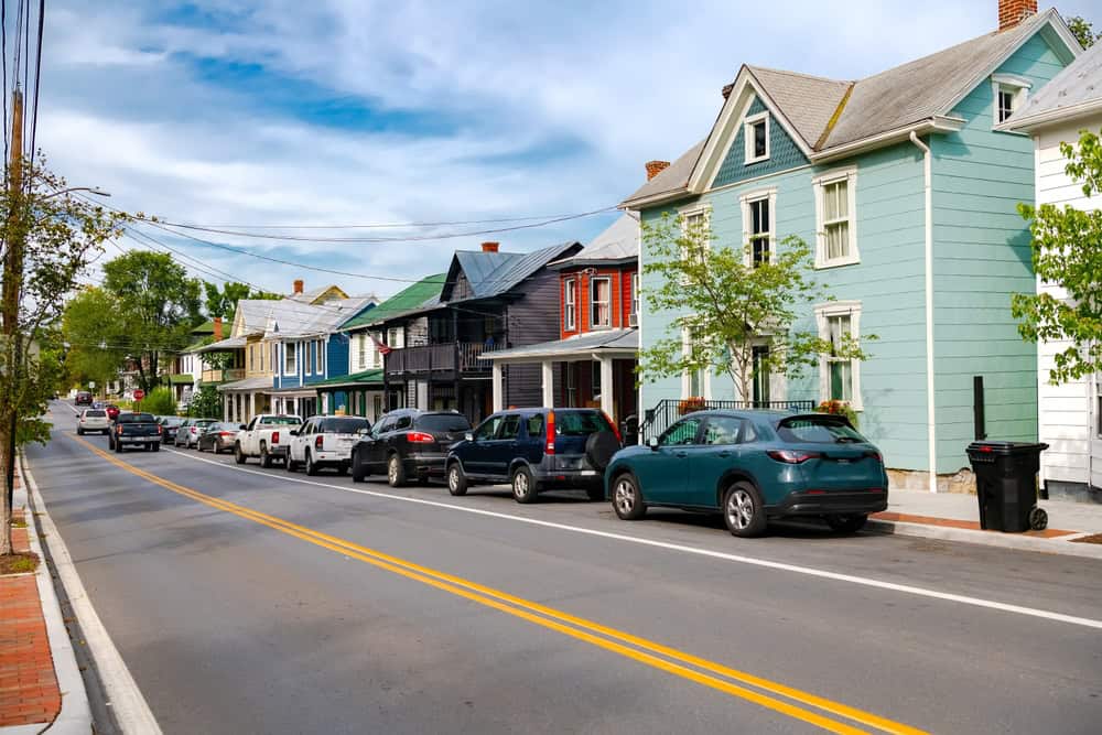 A row of Victorian-era houses in different colors along a two-lane street, with cars parked at the curb in an older town center