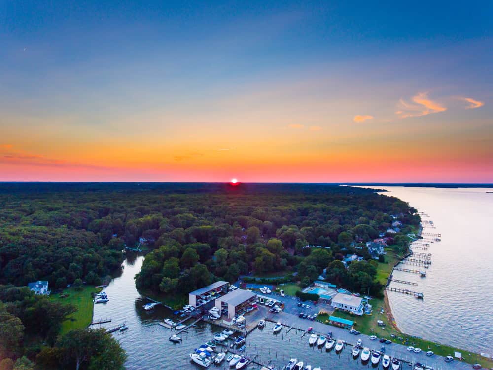 An aerial sunset view of a marina and forested shoreline along the water in Lake Shore, Maryland