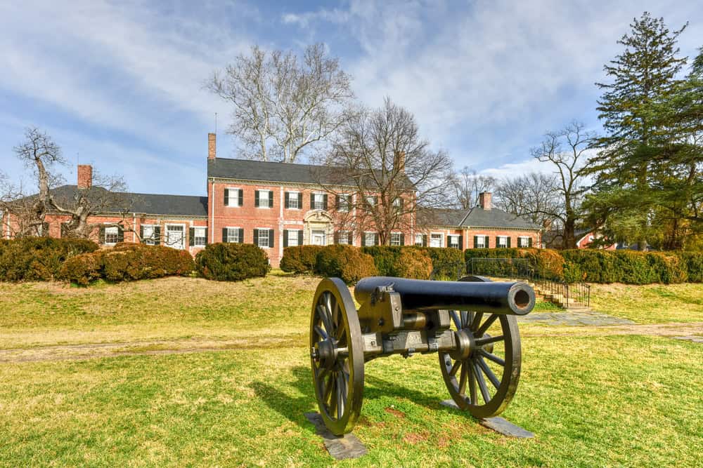 A red brick colonial-era house with a Civil War-era cannon displayed on the lawn, surrounded by bare winter trees