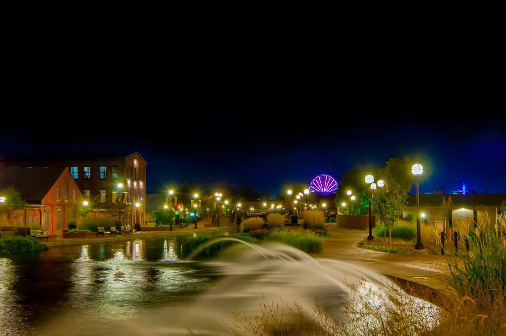 Water flowing over a low spillway beside illuminated buildings at night.