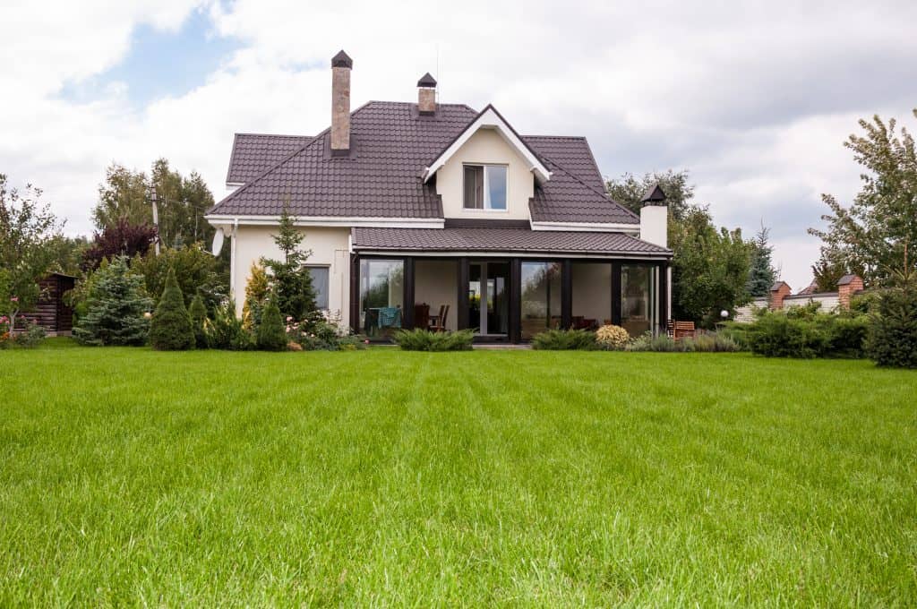 A two-story stucco house with a steep tiled hip roof, two chimneys and a glassed-in porch, behind a wide open lawn edged with evergreen trees