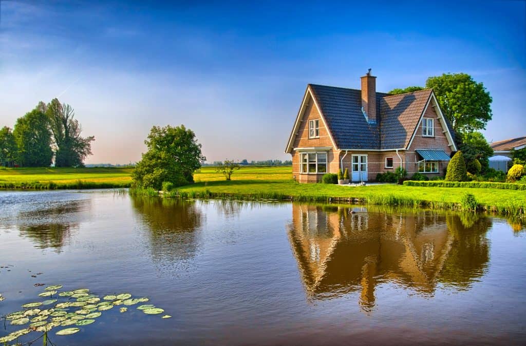 A brick house with a steep tiled roof and chimney beside a still pond, reflected in the water, with open fields and trees around it