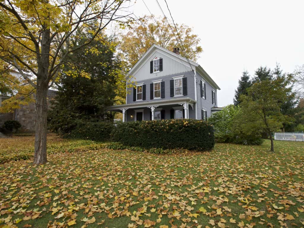 A gray two story Victorian home behind a tall hedge, with autumn leaves covering the lawn in New Franklin, Pennsylvania